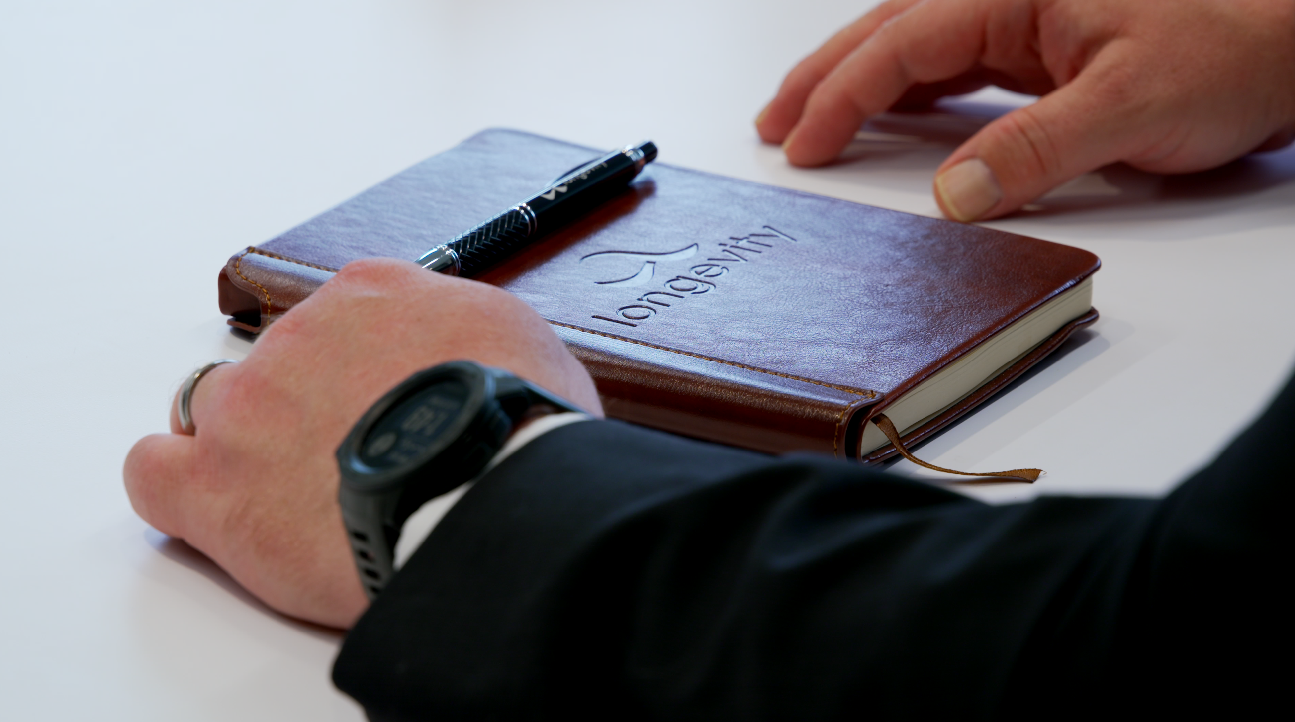 Notebook_FX9 Notebook with the longevity name on the cover sitting on a table. Pen resting on top.
